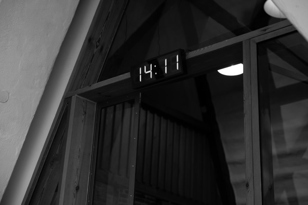 Eine Digitaluhr, die 14:11 Uhr anzeigt, ist über einer Glastür in einem Gebäude mit Holzbalken angebracht, das in Schwarz und Weiß gehalten ist - eine ruhige Umgebung, die man oft bei Exerzitien zum Thema Fasten und Fastenkuren findet.