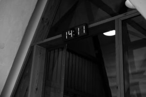 Eine Digitaluhr, die 14:11 Uhr anzeigt, ist über einer Glastür in einem Gebäude mit Holzbalken angebracht, das in Schwarz und Weiß gehalten ist - eine ruhige Umgebung, die man oft bei Exerzitien zum Thema Fasten und Fastenkuren findet.