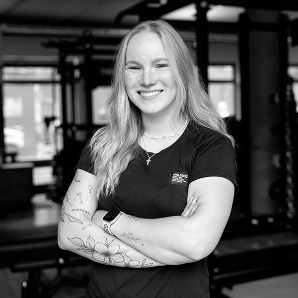 Eine lächelnde Frau mit langen, hellen Haaren steht mit verschränkten Armen in der Turnhalle des Studio St. Pauli und zeigt mehrere Tattoos auf ihren Unterarmen. Sie trägt eine Smartwatch, eine Halskette und ein dunkles T-Shirt. Im Hintergrund sind Fitnessgeräte zu sehen.