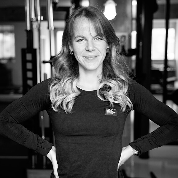 Eine Frau mit gewelltem Haar und langärmeligem Hemd steht selbstbewusst im Studio Sternschanze und lächelt in die Kamera, die Hände in die Hüften gestemmt. Schwarz-Weiß-Bild mit unscharfen Fitnessgeräten im Hintergrund.